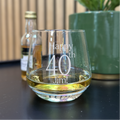 Glass with 'Happy 40th Jim!' engraving, bottle, and plant in the background