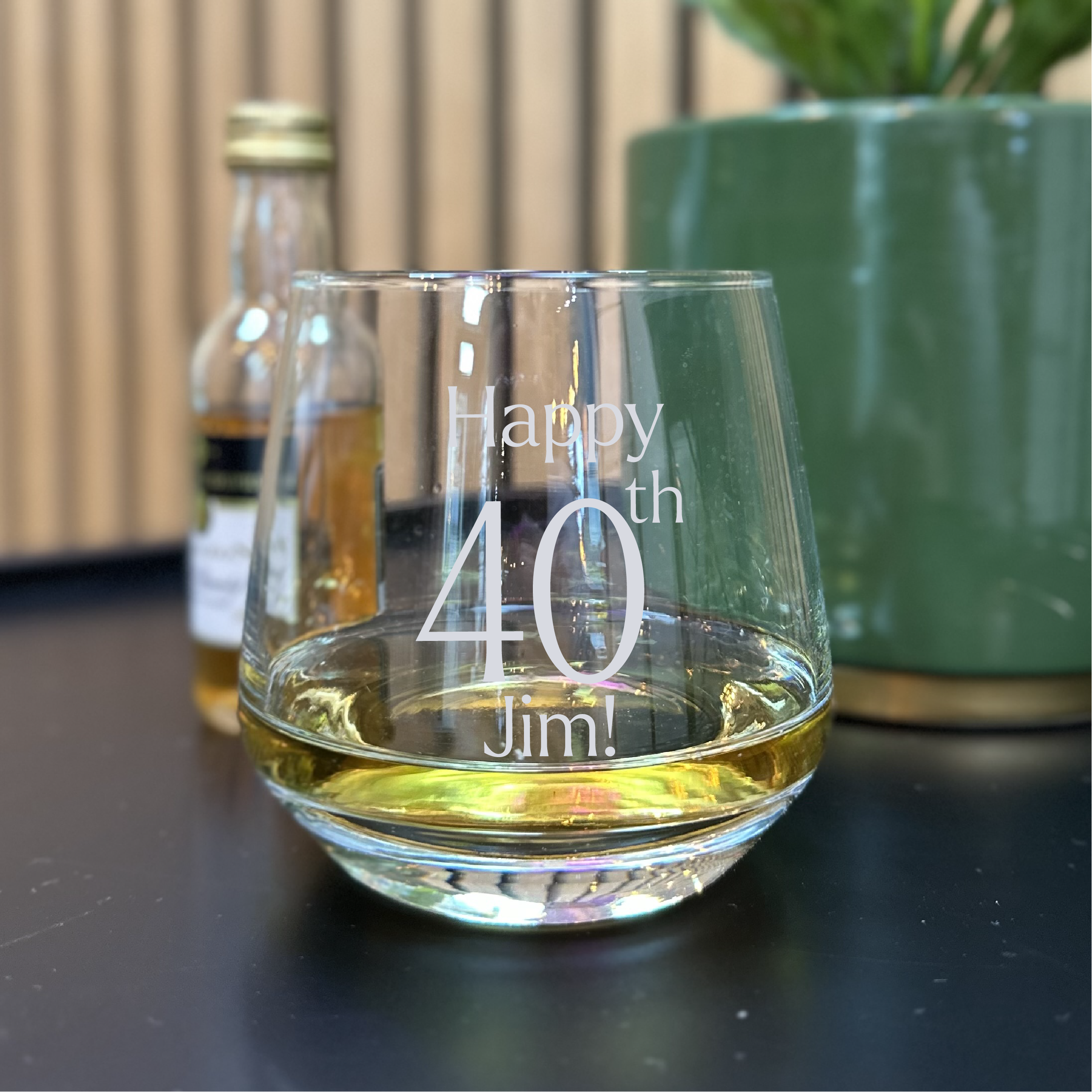 Glass with 'Happy 40th Jim!' engraving, bottle, and plant in the background
