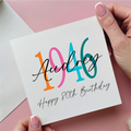 Hand holding a card with 'Audrey 1946 Happy 80th Birthday' text on a pink background