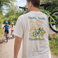 Man wearing a 'Daddy Smith' t-shirt with a bicycle graphic and text, standing outdoors with children on bikes in the background.