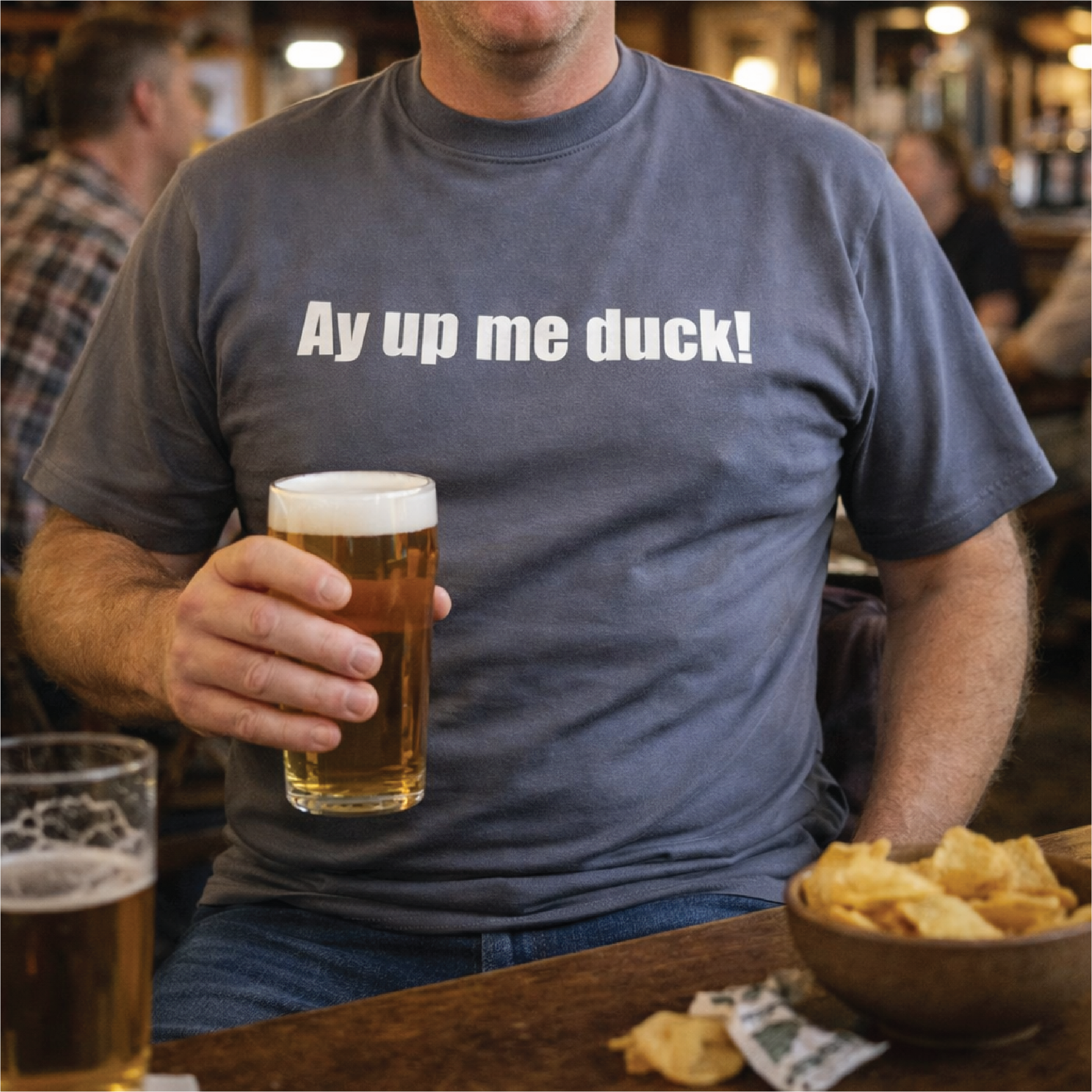 Man wearing a t-shirt with 'Ay up me duck!' text, holding a beer in a bar setting.