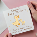 Baby shower card with ducks and text held by a hand on a pink background