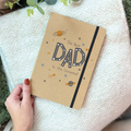 Notebook with 'The Best Dad in the Universe' text held by a hand on a textured surface.