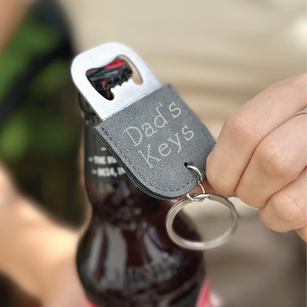 Bottle opener keychain with engraved text held by a hand over a bottle.