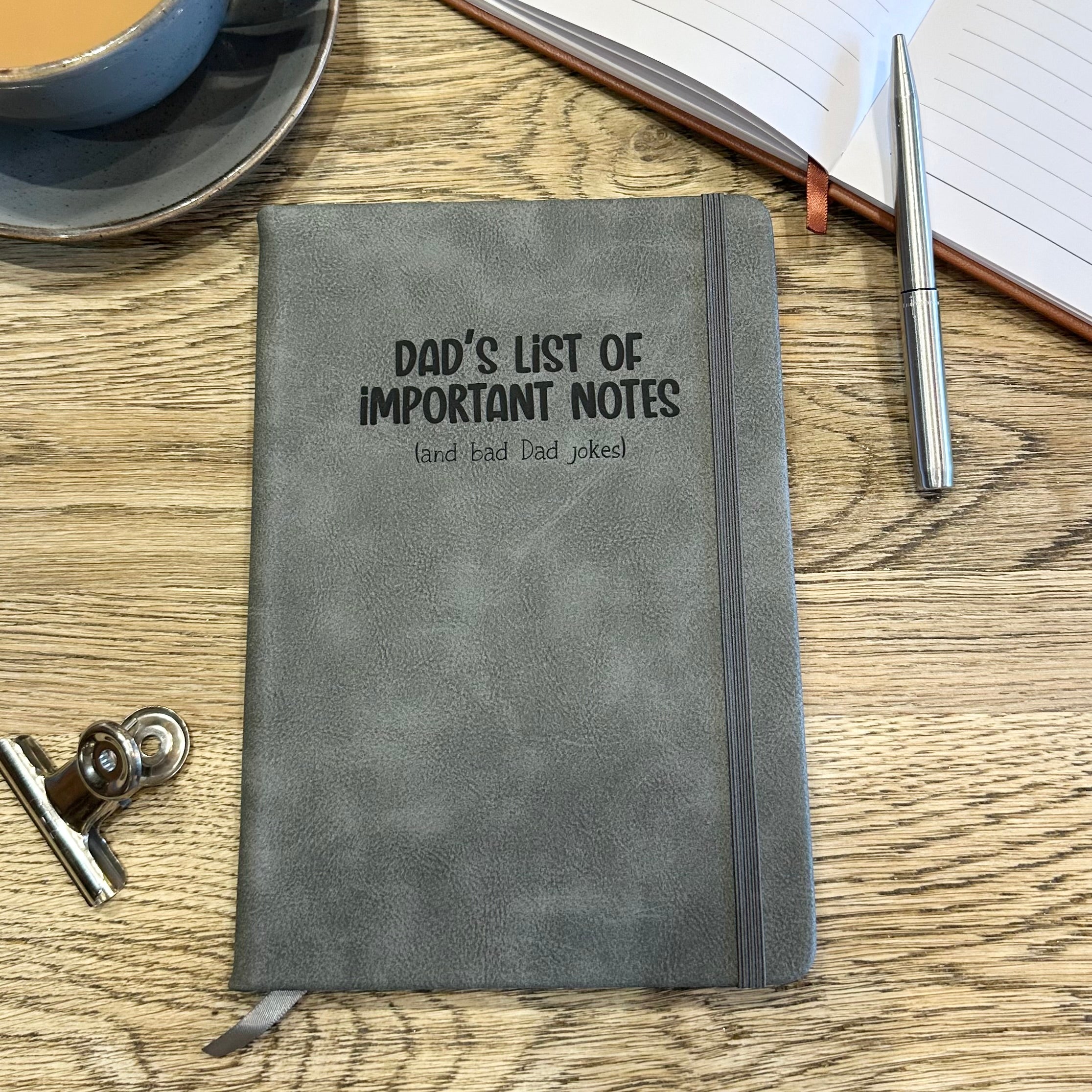Gray notebook with 'Dad's List of Important Notes' text on a wooden table with a cup and pen.