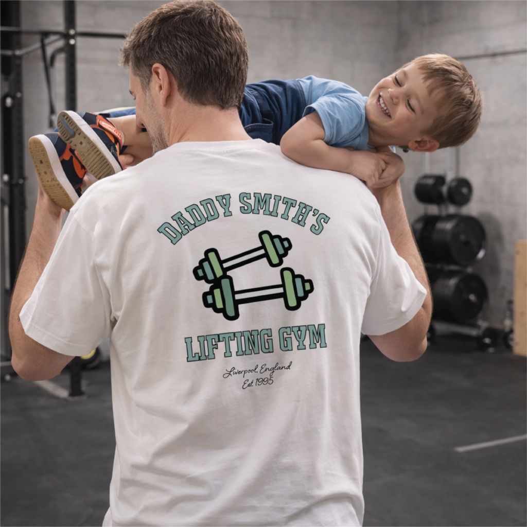 Man lifting a child on his shoulders with a 'Daddy Smith's Lifting Gym' t-shirt in a gym setting.