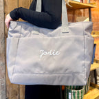 Person holding a gray tote bag with 'Jodie' embroidered on it, outdoors.