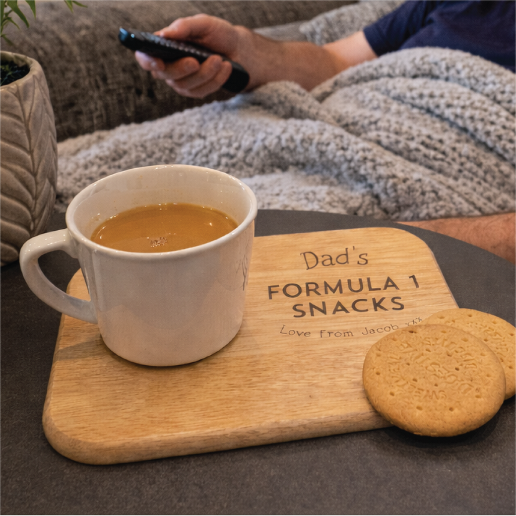 Person watching TV with a mug of coffee and cookies on a tray labeled 'Dad's Formula 1 Snacks'.