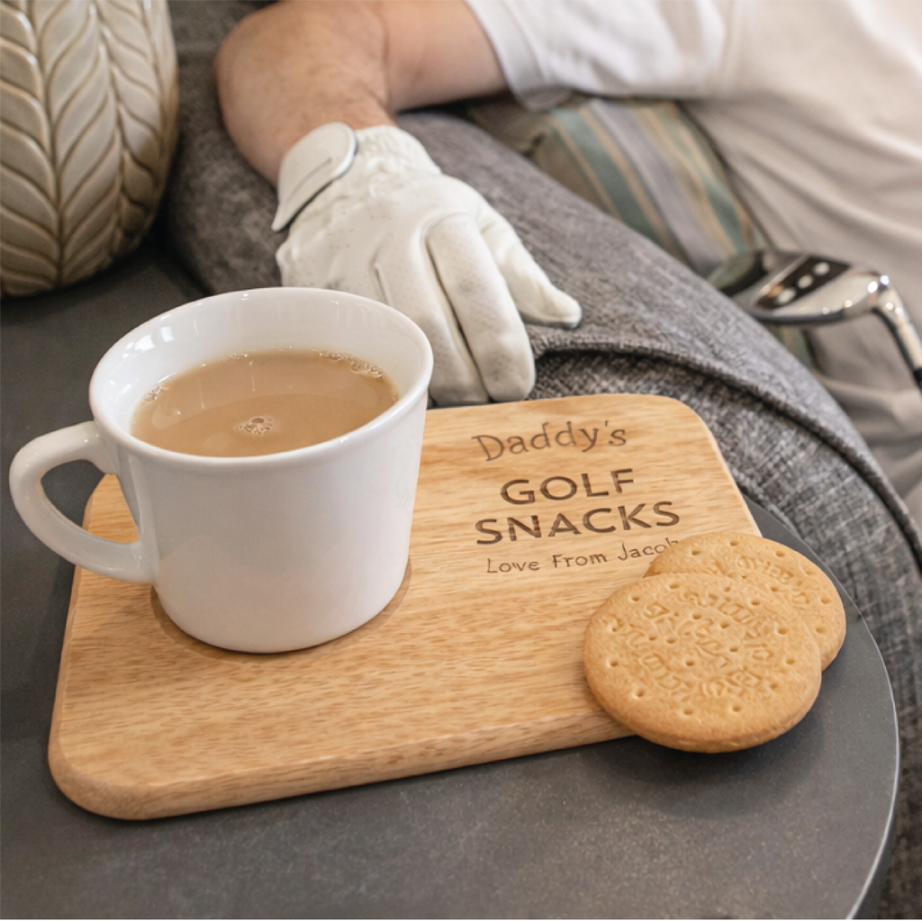 White mug with coffee and a cookie on a wooden board with 'Daddy's Golf Snacks' text, person wearing white glove in background.