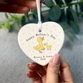 Heart-shaped 'Happy Mother's Day' ornament with ducks held by a hand against a blurred green leaf background.