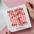 Card with 'My Mum, My Hero' text and illustration held by a hand on a pink background