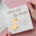 Card with 'Welcome to the World' text and a duck illustration, held by a hand on a pink background.