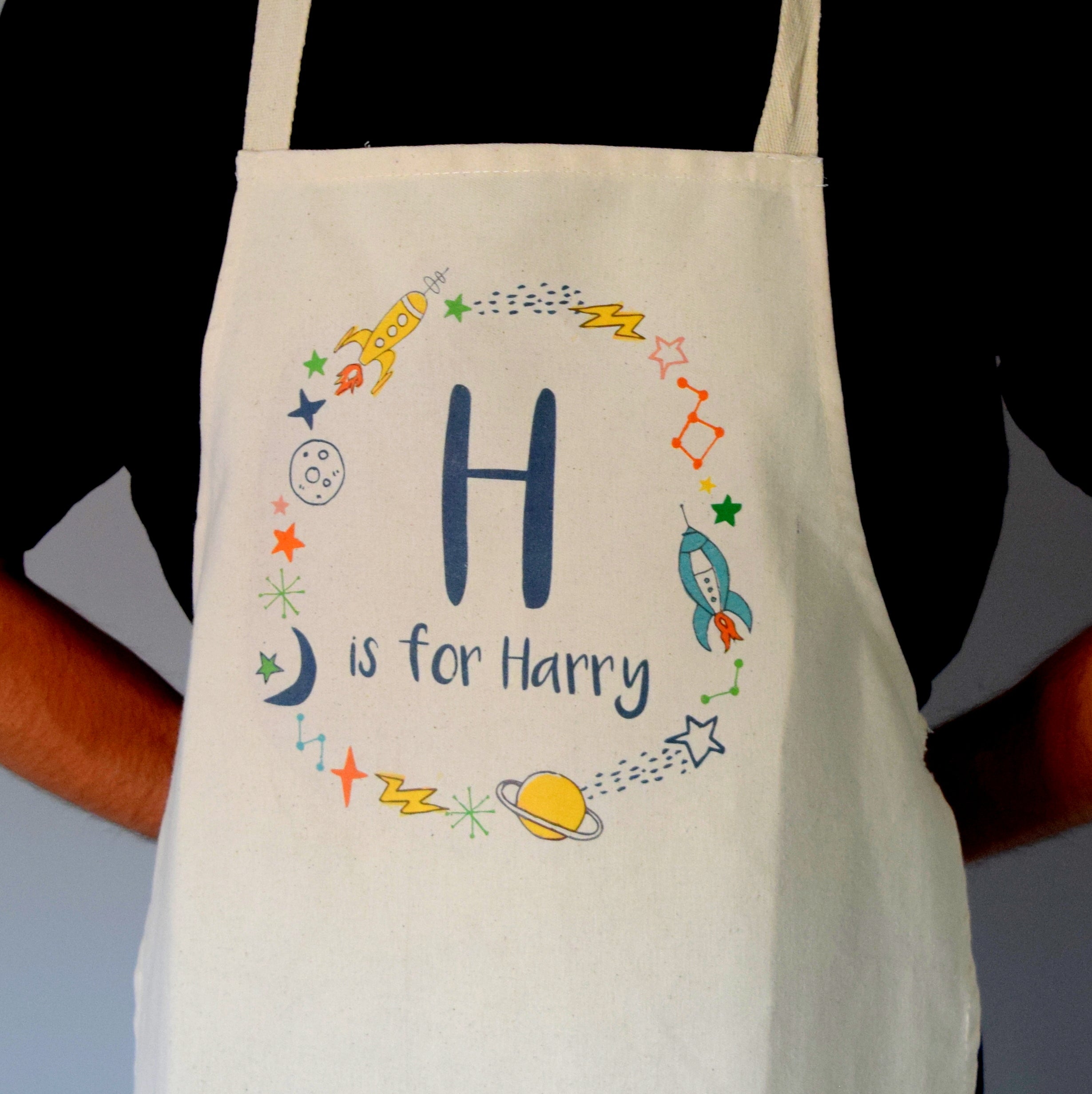Beige apron with 'H is for Harry' text and space-themed graphics on a black background.