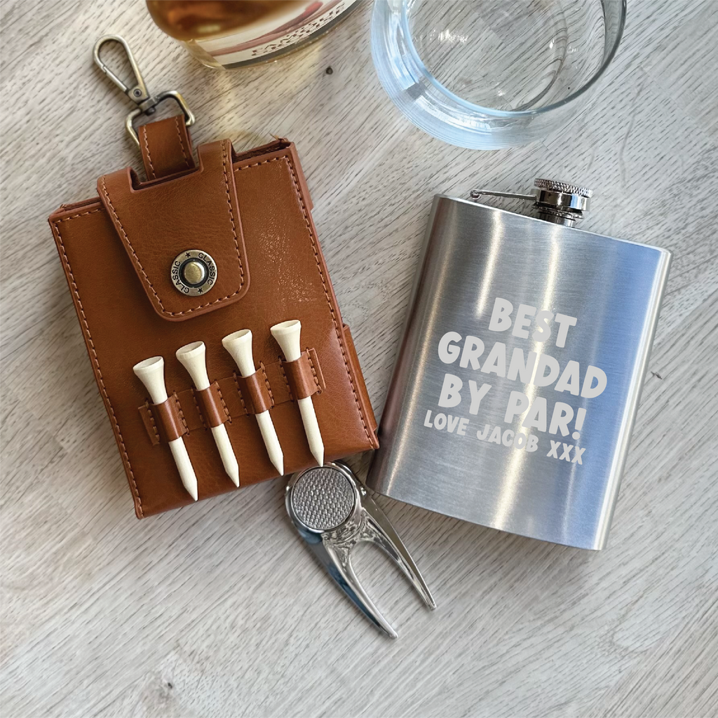 Golf-themed items including a leather tees holder, golf tees, divot tool, and a silver flask with engraved text on a wooden surface.