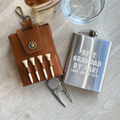 Golf-themed items including a leather tees holder, golf tees, divot tool, and a silver flask with engraved text on a wooden surface.