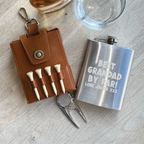 Golf-themed items including a leather tees holder, golf tees, divot tool, and a silver flask with engraved text on a wooden surface.