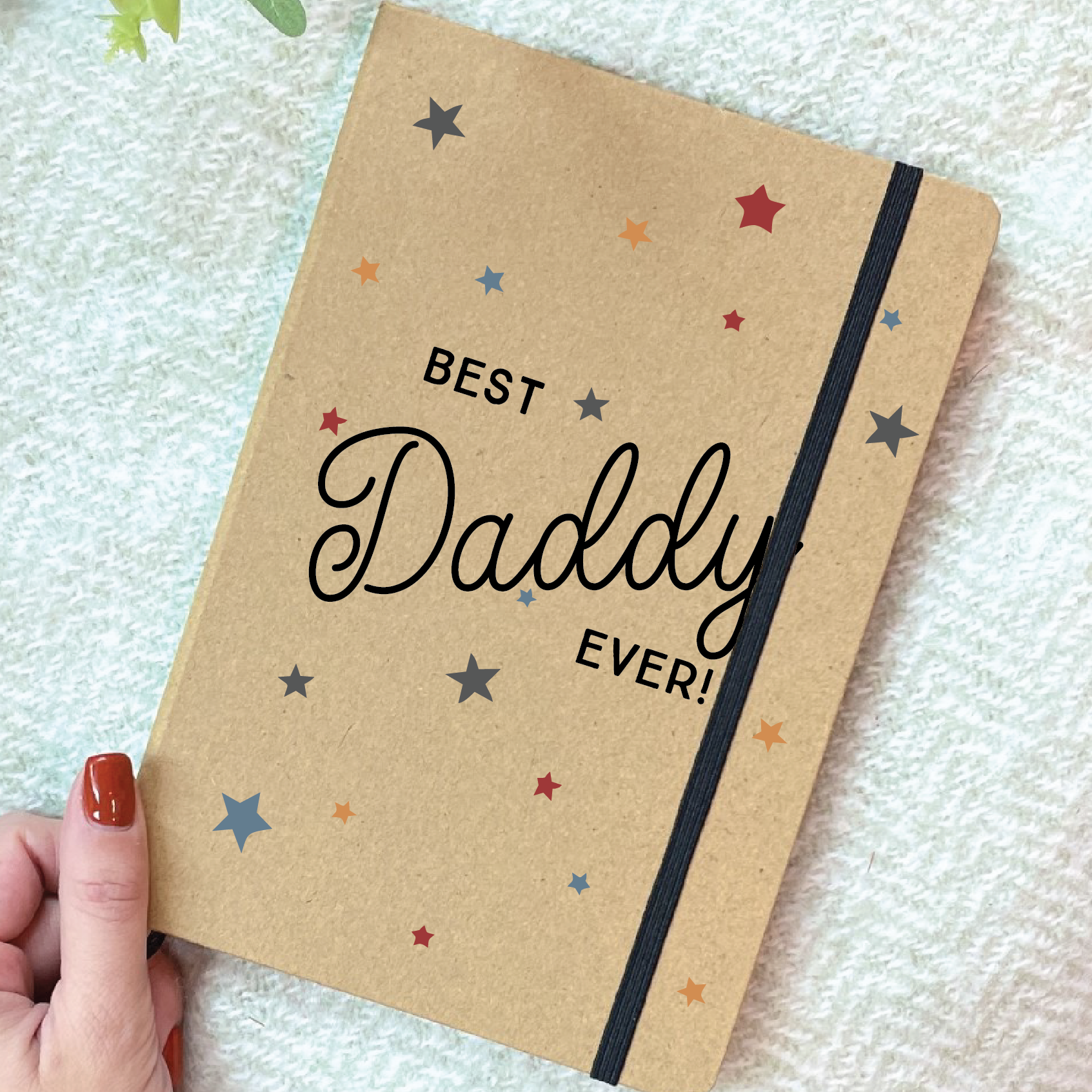 Notebook with 'Best Daddy Ever!' text held by a hand on a textured surface.