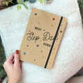 Notebook with 'Best Step Dad Ever!' text held by a hand on a textured surface.