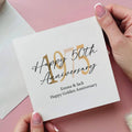 Hand holding a card with 'Happy 50th Anniversary' and '1975' text on a pink background