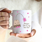 Person holding a mug with a cartoon tea bag design and text, wearing a white sweater.