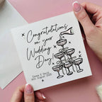 Hand holding a wedding card with 'Congratulations on your Wedding Day!' text and wine glass illustrations.
