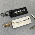 Two keychains on a gray textured background, one with 'Drive Safe' message and the other with 'You've Passed' message.