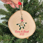 Wooden ornament with mistletoe design and 'Emma & David 2023' text, hanging on a Christmas tree.