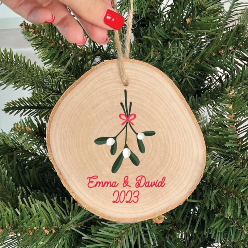 Wooden ornament with mistletoe design and 'Emma & David 2023' text, hanging on a Christmas tree.