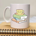 White mug with a design of books and a teacup on top, placed on a wooden surface.