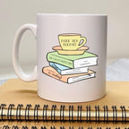 White mug with a design of books and a teacup on top, placed on a wooden surface.