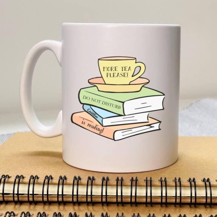 White mug with a design of books and a teacup on top, placed on a wooden surface.