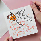 Card with an illustration of a cherub holding a cocktail and text, held by hands on a pink background.