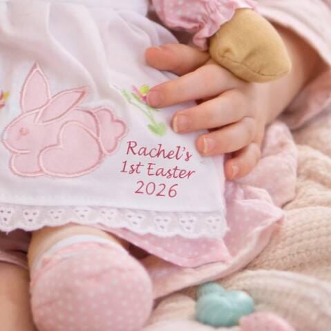 Baby's outfit with embroidered bunny and text 'Rachel's 1st Easter 2026' held by a child's hand.