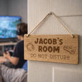 Wooden sign on a door with 'Jacob's Room Do Not Disturb' text, person in the background using a computer.