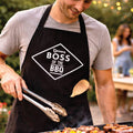 Man wearing a black apron with 'Boss of the BBQ' text, grilling outdoors.