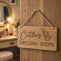 Wooden sign hanging on a door with 'Caitlin's Dressing Room' engraved, in a vanity area.