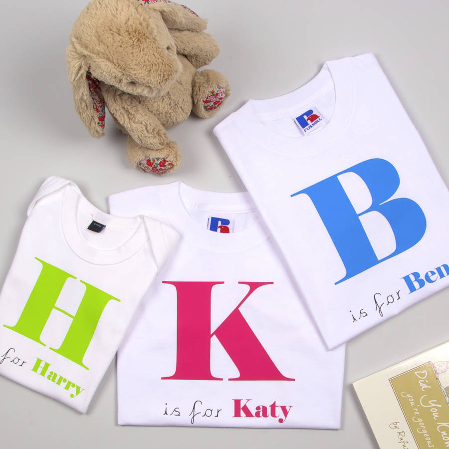 Three white t-shirts with colorful letters and names on a light gray surface with a teddy bear.