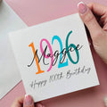 Hand holding a card with '1926 Maggie Happy 100th Birthday' text on a pink background