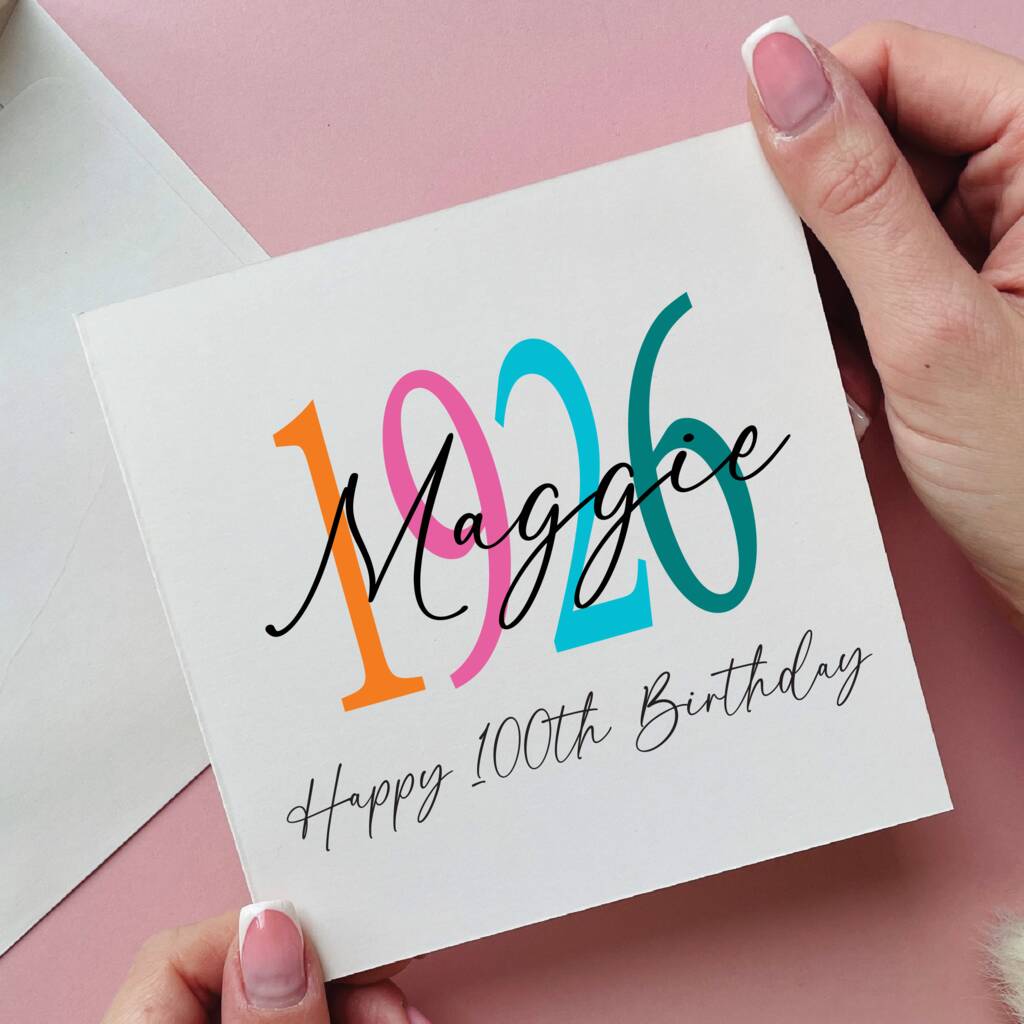 Hand holding a card with '1926 Maggie Happy 100th Birthday' text on a pink background