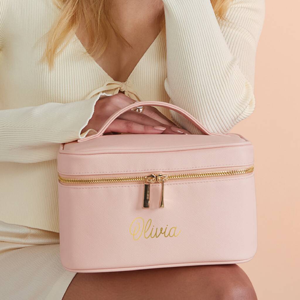 Pink cosmetic bag with gold zipper and 'Olivia' engraving held by a person in a cream outfit.