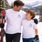 Man and child in matching white t-shirts with a logo, standing in a snowy landscape with skis.