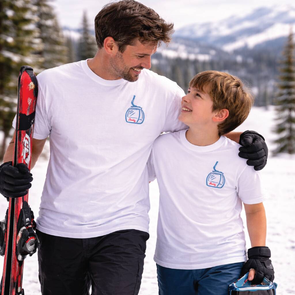 Man and child in matching white t-shirts with a logo, standing in a snowy landscape with skis.