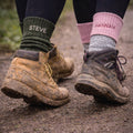 Two pairs of feet wearing muddy boots and personalized socks on a natural ground.