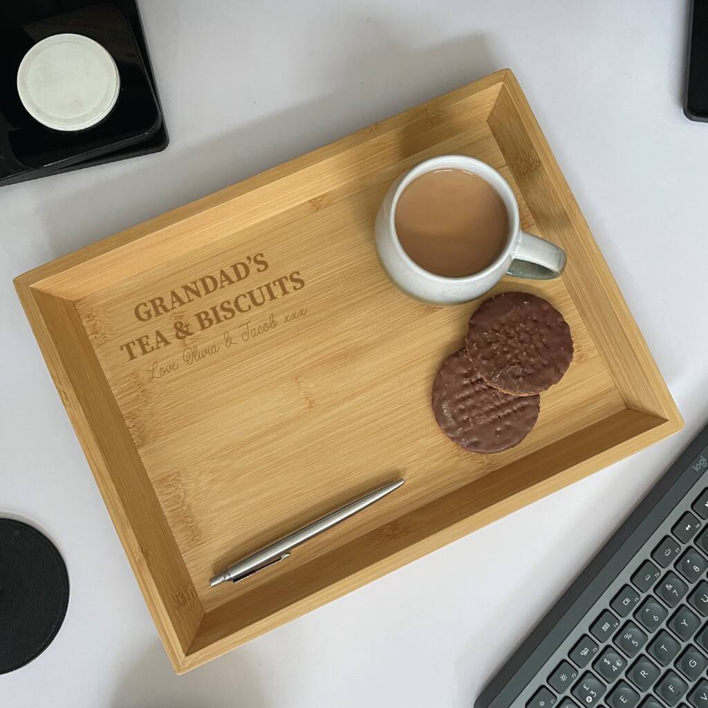 Wooden tray with 'Grandad's Tea & Biscuits' engraving, cup of tea, cookies, and pen on a white surface.